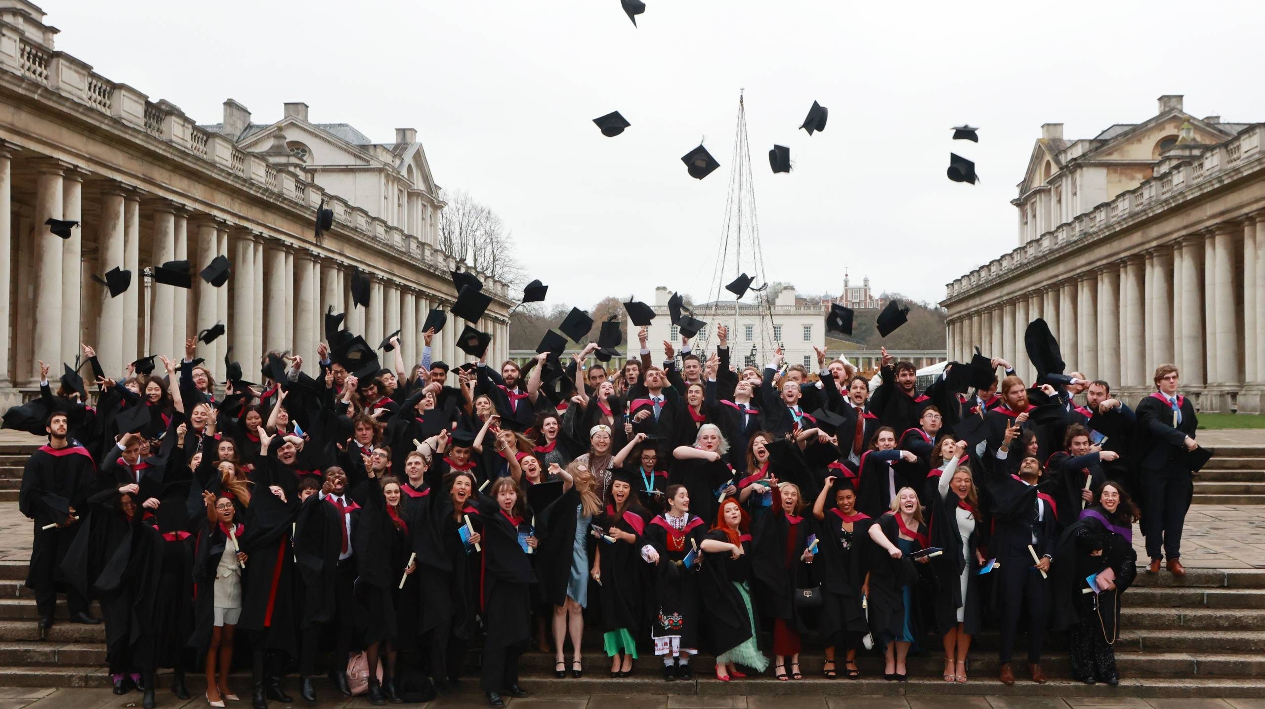 A group of students in academic robes throw their hats into the air in celebration between columned buildings