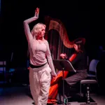 A dancer dressed in white lifts her right hand above her head in front of a musician in a black costume who plays the harp.