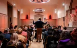 A conductor, with his back facing an audience, leads a band of instrumentalists in a grand practice room with red lighting.