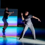 Two dancers dressed in black move against a multi-coloured background in the centre of a stage.