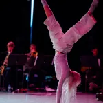 A dancer in white does a handstand in pink lighting, with instrumentalists performing in the backdrop.
