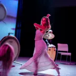 A dancer in a white top and skirt steps out on stage in pink lighting.