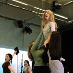 A student with long blonde hair is lifted up into the air by two fellow students. Two dancers glide by on the left.