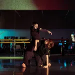 Two dancers move centre stage in a studio. A cellist plays on the left and a flautist performs on the right.