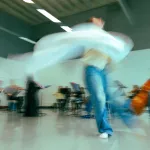A dancer spins in the centre of a brightly-lit dance studio, her white costume spins against the backdrop of musicians.