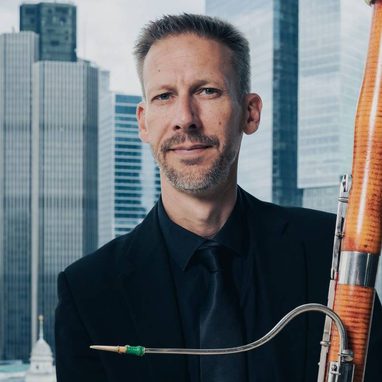 Graham Hobbs is a white man with short hair. He is wearing a dark coloured suit and holding a bassoon.