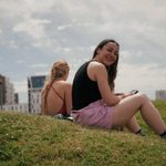 Two students sit on a hill overlooking the Laban Building. One students wearing a black vest top and pink shorts turns to look at the camera.