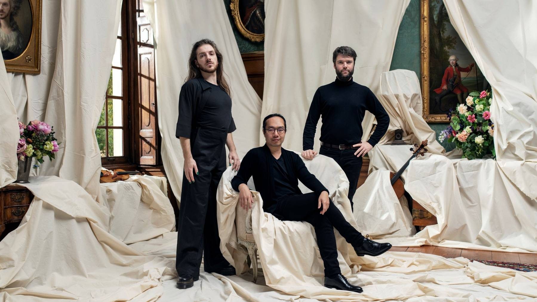 Linos Piano Trio, photo shoot by Tim Mintiens, the trio stand in a drawing room with white sheets draped across the furniture, and white curtains billowing from open windows