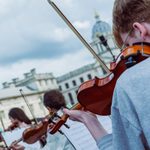 Young Musicians Summer School, violin players follow their sheet music with the old royal naval college and a cloud sky behind them