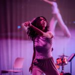 Dancer in purple lighting with her left hand outstretched in front of her. The background screen imitates her movement.