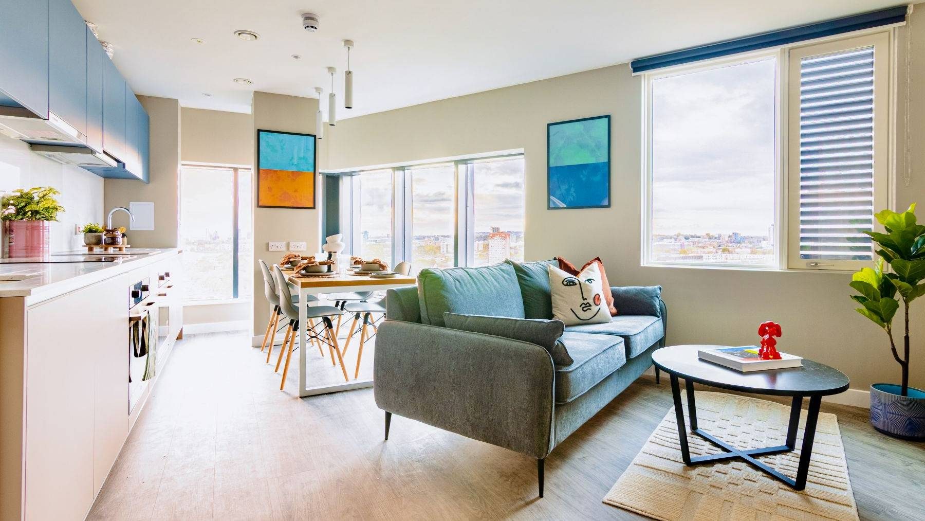 Shared kitchen with a view across the city from the windows. Within the room are a teal sofa in front of a circular coffee table. Behind, is a shared dining table and kitchen facilities.