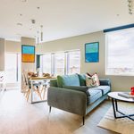Shared kitchen with a view across the city from the windows. Within the room are a teal sofa in front of a circular coffee table. Behind, is a shared dining table and kitchen facilities.