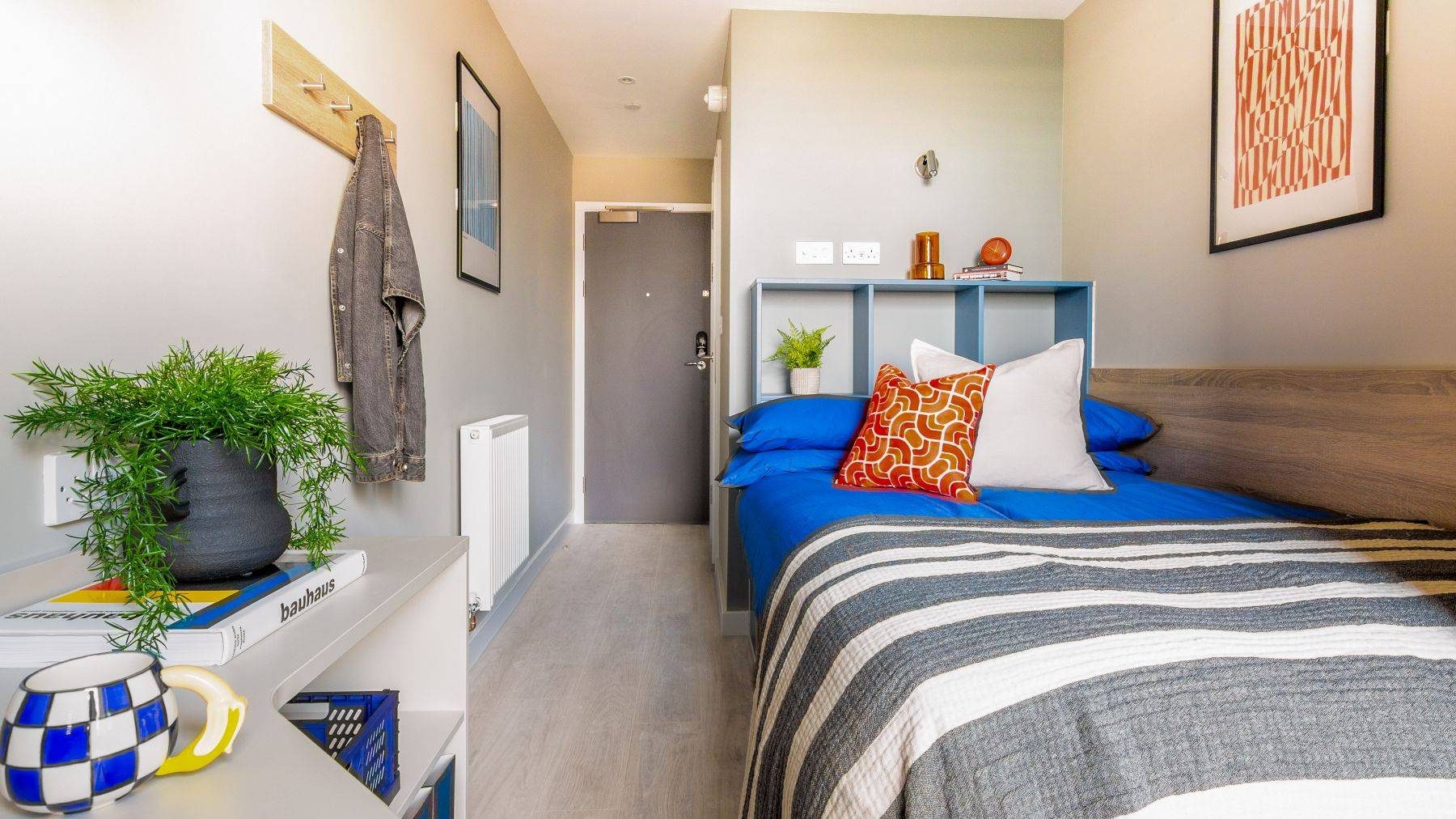 Ironworks standard ensuite bedroom, small double bed with cubby holes for storage above. To the side of the bed is a desk with bookshelving. A jacket is hung on pegs along the wall alongside bright modern art in frames.