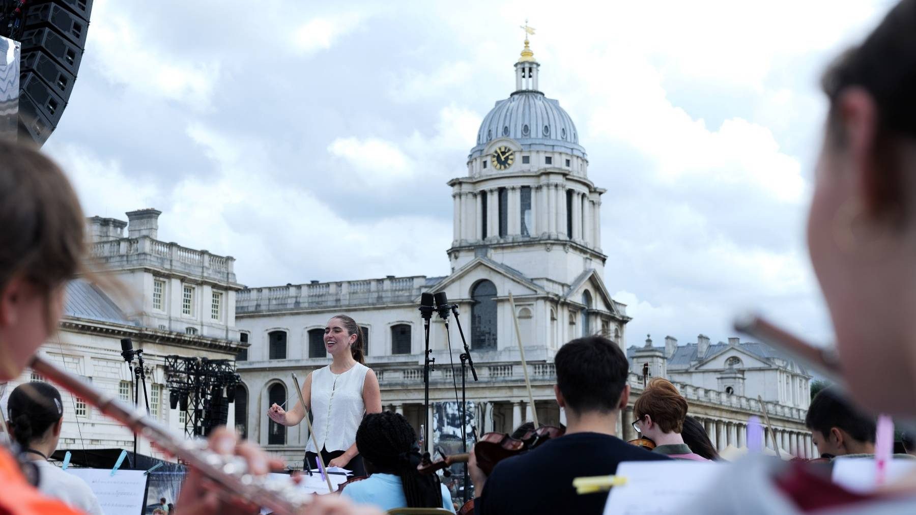 Students playing woodwind and strings instruments with a smiling conductor, on stage at the Old Royal Naval College in London