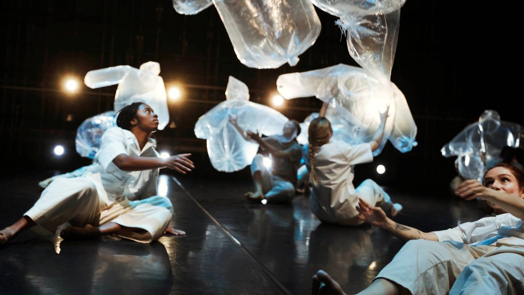 Postgraduate Dance Performance students perform a dance titled 'Clouds', stage lights illuminate four performers in seated and reclining positions while inflated transparent plastic ponchos float above them ethereally.