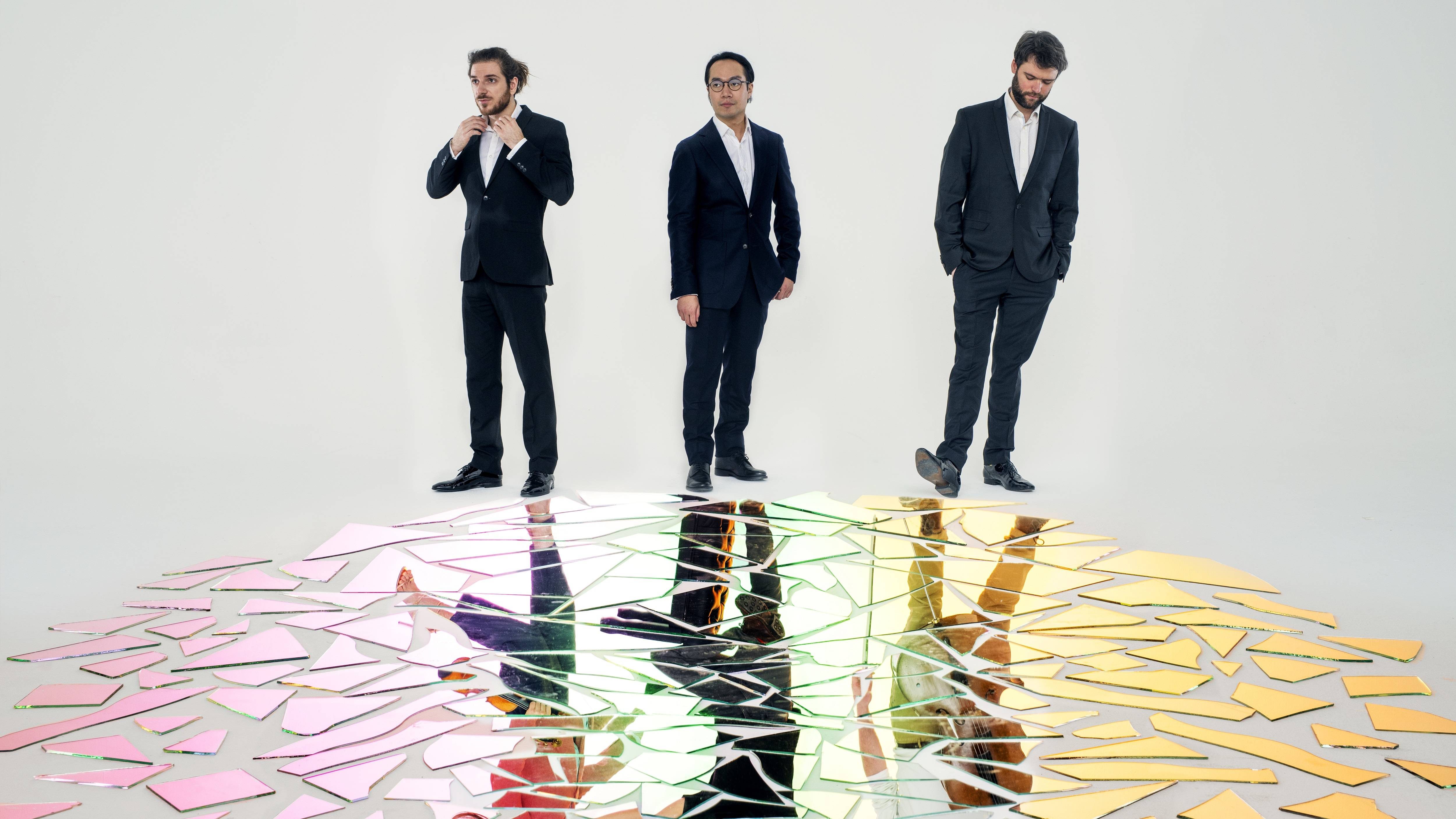 The Linos Piano Trio in smart suits, holding their instruments, with dynamic reflections in coloured glass at their feet.