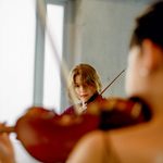 Two students playing violin, one in foreground out of focus, a second in the background looking at the camera