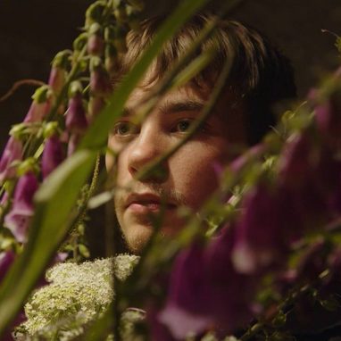 Portrait of Samuel D Loveless looking through purple foxgloves and green foliage, with his face partially obscured by the plants. Warm, soft lighting against a dark background.